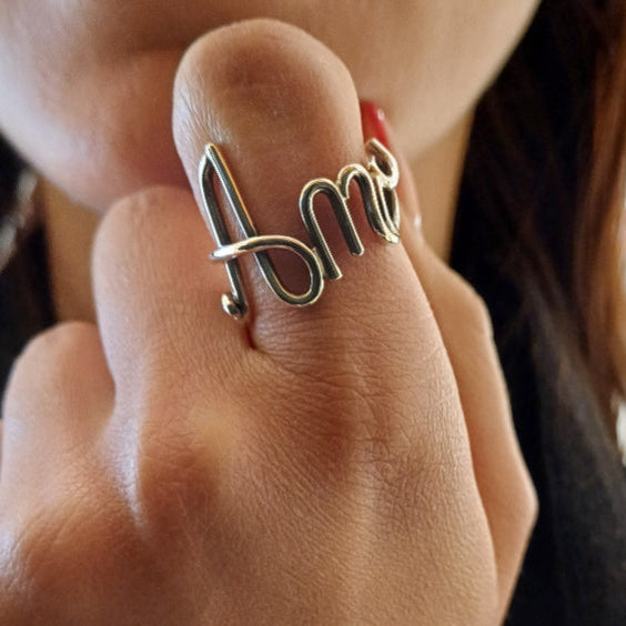 Immagina di accarezzare un filo d’argento liscio e brillante, che si snoda e si intreccia con eleganza fino a formare un nome. È un anello leggero e morbido al tatto, con curve armoniose e una struttura aperta, che segue dolcemente la forma del dito. Ogni lettera è parte di un unico filo, senza interruzioni, donando una sensazione di fluidità e continuità. Un gioiello su misura, pieno di emozione e personalità.