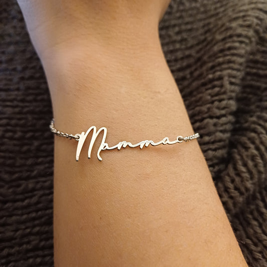 Bracciale sottile e leggero in argento 925, con catena a maglia fine e chiusura ad anello a molla. Al centro, una scritta liscia e lucida in corsivo: "Mamma", tagliata in un unico pezzo di metallo, con la lettera “M” leggermente più alta e curva. Il metallo è liscio al tatto, senza spigoli, con una finitura brillante. Il bracciale è morbido al polso e pensato per un uso quotidiano, leggero e confortevole.