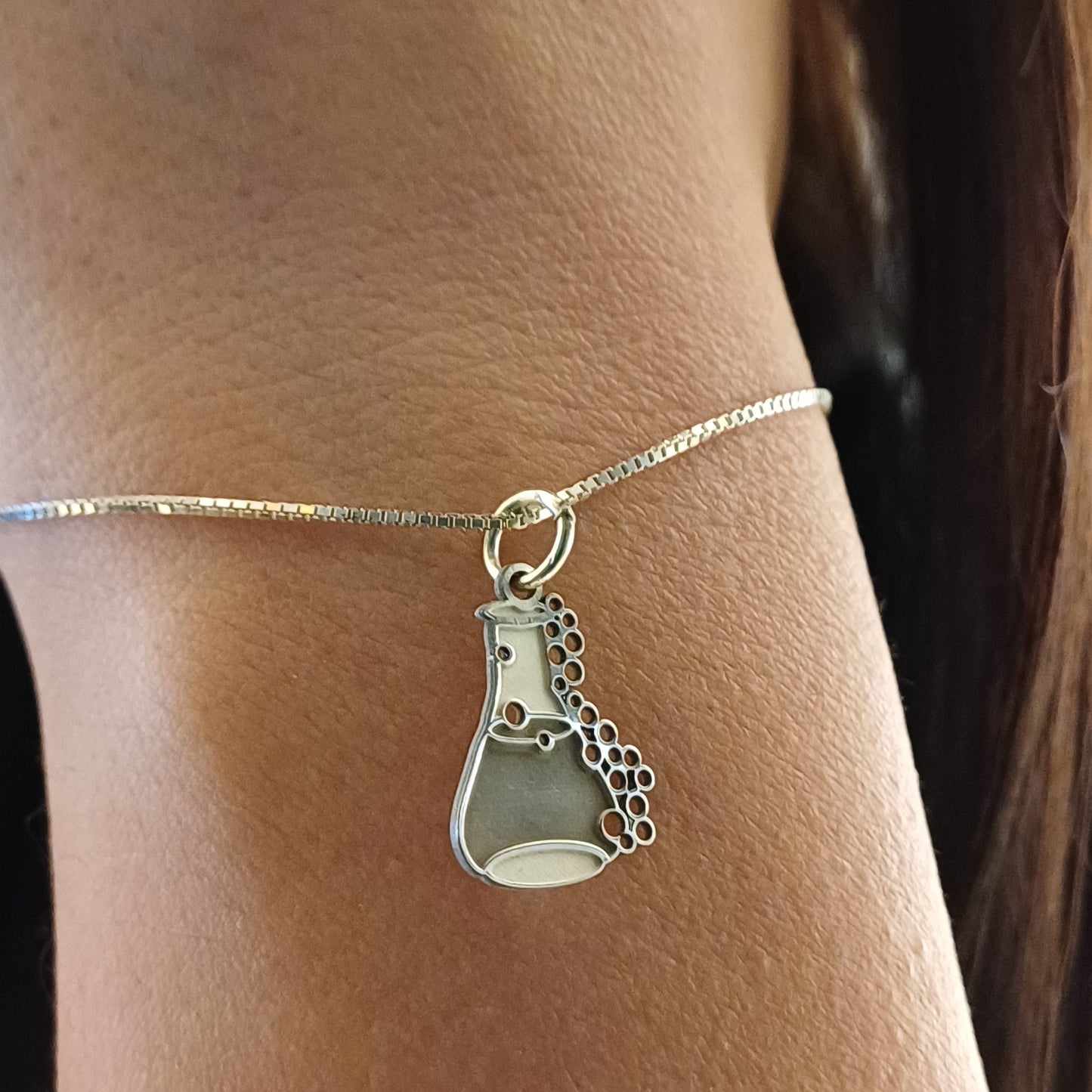 Il ciondolo rappresenta una piccola ampolla da laboratorio, alta circa 1,3 cm, realizzata in argento 925. La superficie è liscia e satinata, con rilievi che rappresentano delle bollicine che sembrano salire lungo il bordo. L’oggetto è leggero, con forme morbide e linee dolci. Al tatto, le bolle e il bordo dell’ampolla offrono una piacevole variazione tra pieno e vuoto, rendendolo facile da riconoscere e molto evocativo.