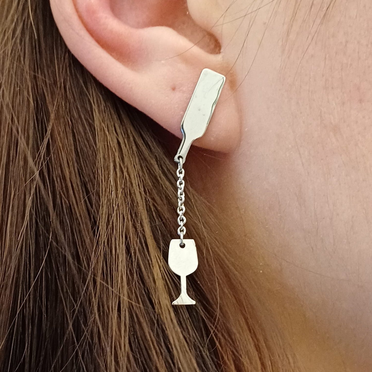 Questi orecchini pendenti in argento 925 sono composti da due elementi collegati da una sottile catenella. La parte superiore rappresenta il collo di una bottiglia, mentre il ciondolo inferiore raffigura un calice da vino inclinato, come se stesse versando il suo contenuto. Alcune versioni presentano una smaltatura a caldo in un intenso colore rosso vino. La superficie è incisa al laser, con dettagli ben definiti. Leggeri e lunghi 5 cm, si muovono delicatamente seguendo i movimenti del viso.