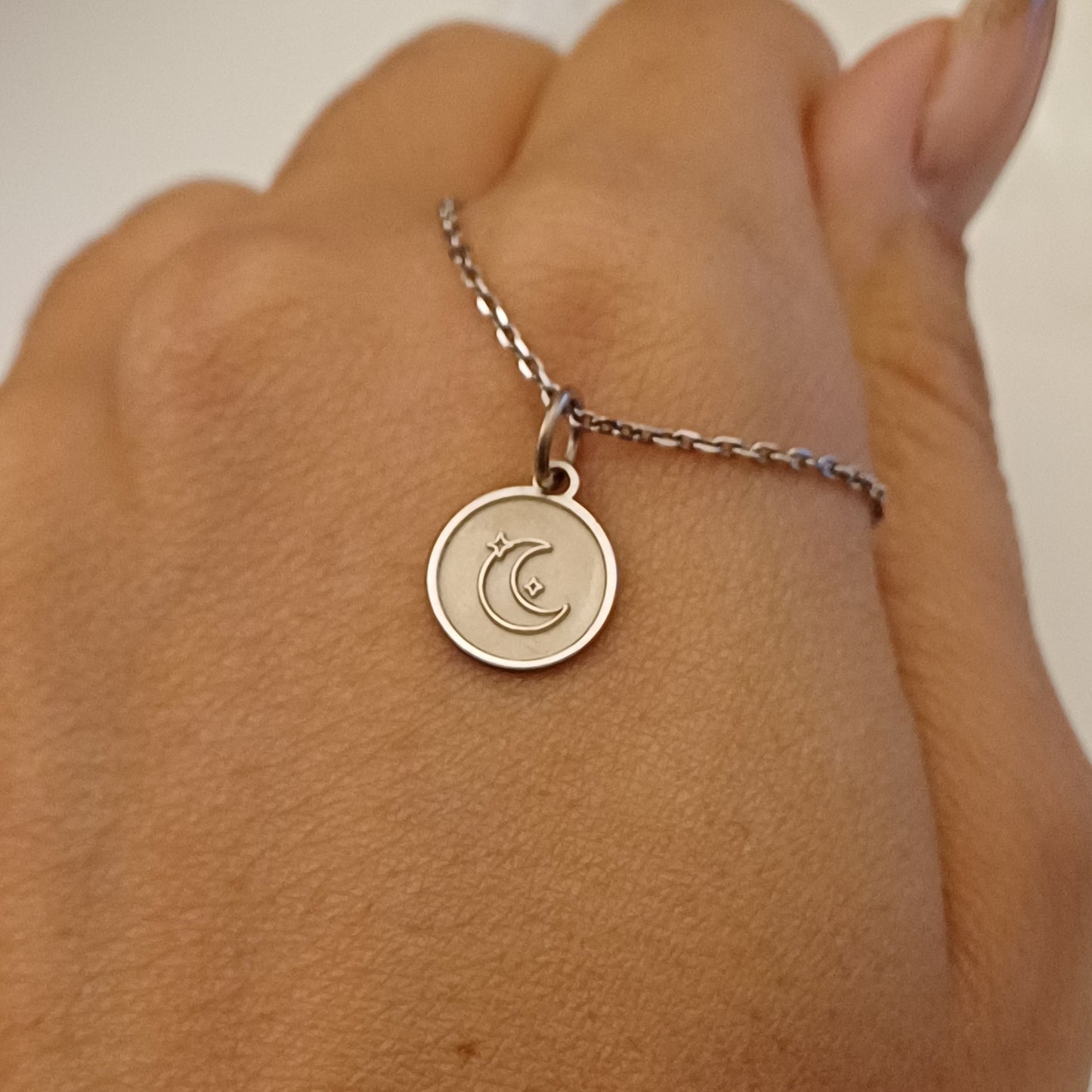 Hai tra le mani una piccola medaglia rotonda in argento, dal diametro di circa un centimetro. La superficie è liscia e satinata, mentre il centro è decorato con una luna crescente in rilievo e due stelline incise. Sul retro, puoi scegliere di far incidere una frase o una dedica. È un gioiello leggero e simbolico, creato a mano in un laboratorio orafo, pensato per accompagnare chi lo indossa in modo discreto e personale.