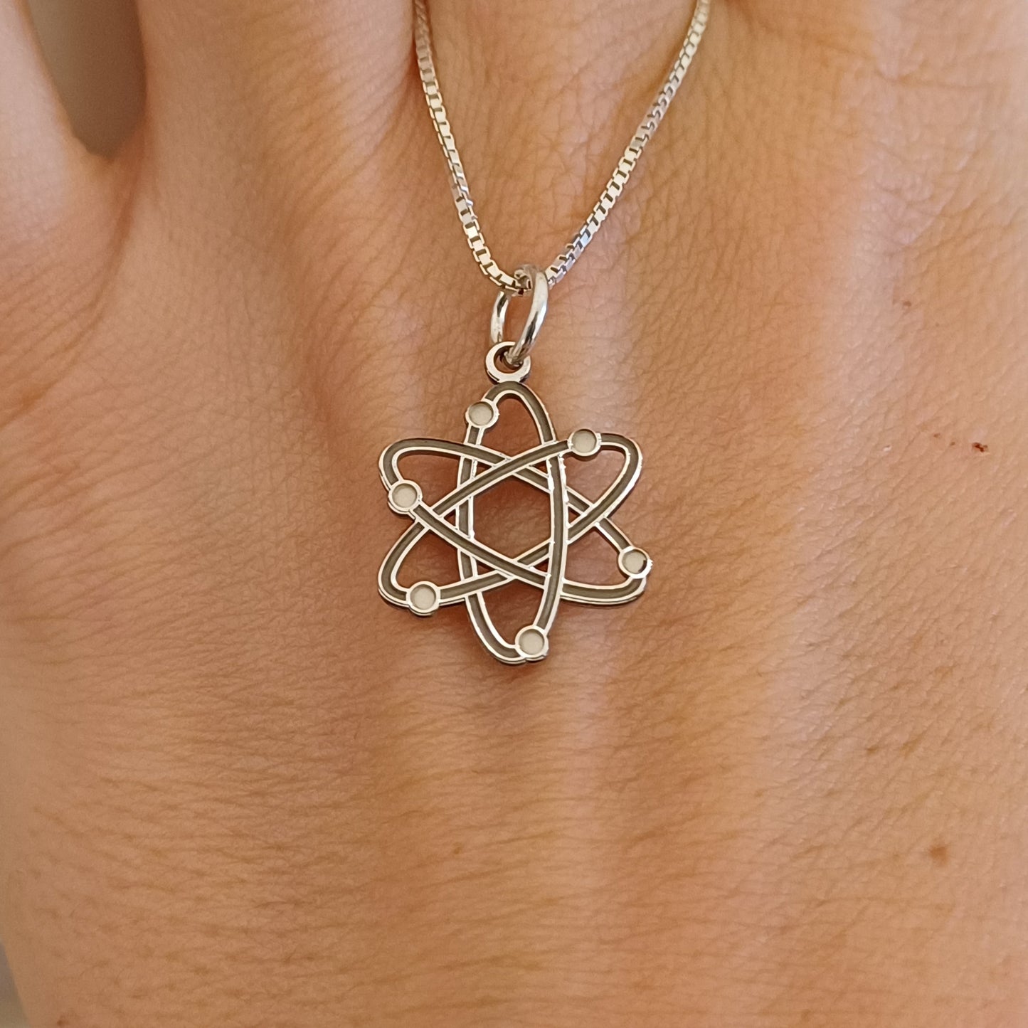Questo ciondolo rappresenta un atomo stilizzato, con sei orbite che si intrecciano creando una forma armoniosa e simmetrica. È in argento 925, con una finitura mista: liscia e satinata. Al tatto, si percepiscono i cerchi morbidi e leggermente bombati e piccole sfere posizionate lungo le orbite. È leggero, misura 1,4 cm di altezza ed è piatto, ma con rilievi netti che lo rendono facile da riconoscere anche solo con le dita.