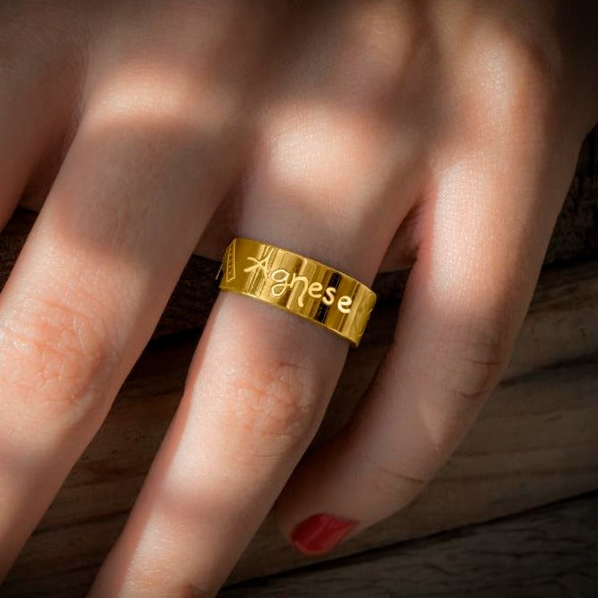L'immagine  della fascia indossata mostra un anello in oro giallo con una superficie liscia e lucida. Sulla fascia dell'anello sono incisi dei simboli e il nome "Agnese" in uno stile di scrittura a mano. Accanto al nome, c'è un piccolo disegno che rappresenta un righello ,un orologio, un peso ed un calendario tutti con incisi i dati della nascita. L'anello ha un design semplice ma elegante, con incisioni ben definite e dettagli che lo rendono unico 