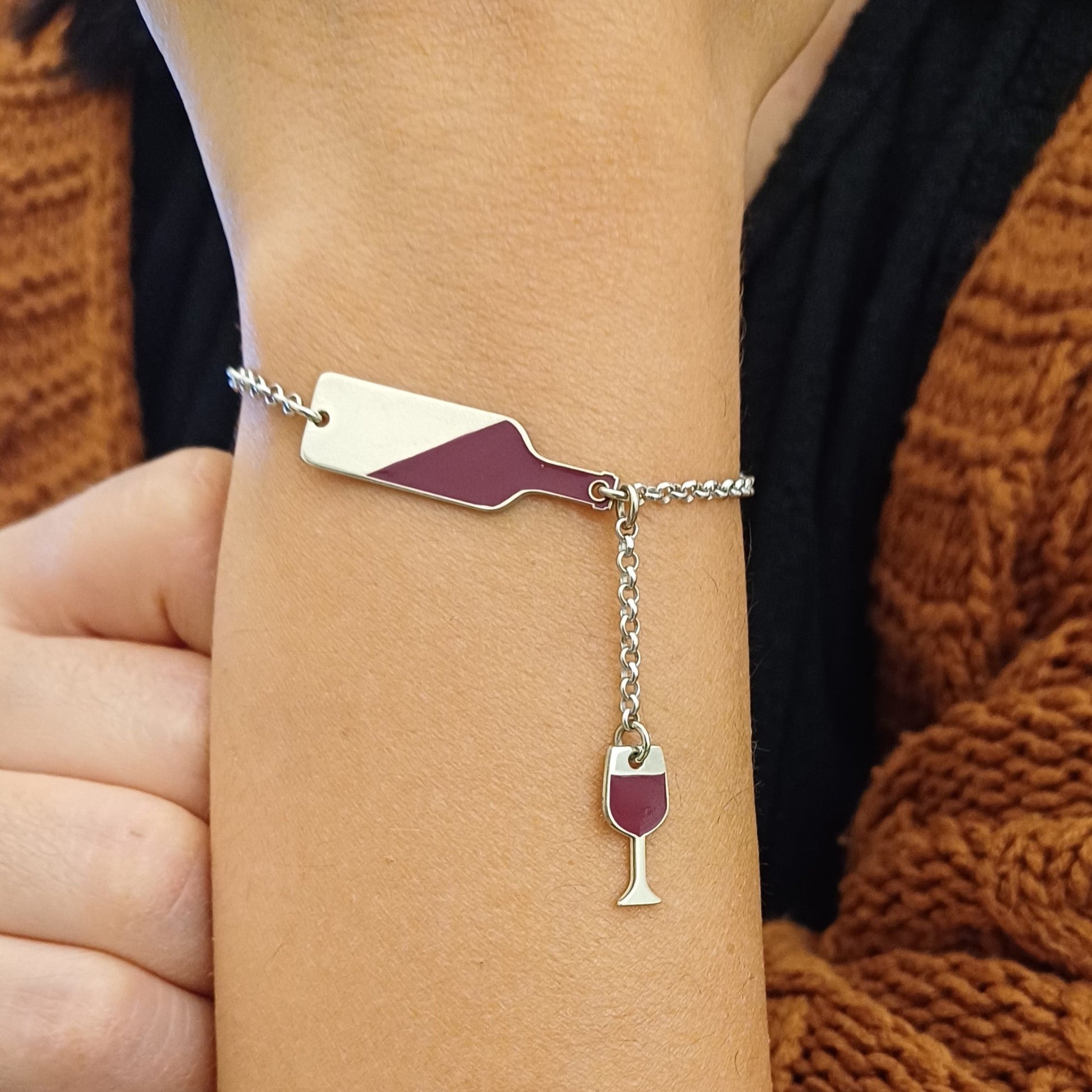  bracciale in argento con un design unico: una bottiglia di vino stilizzata smaltata di rosso, a cui è collegato un piccolo calice pendente. La bottiglia, smaltata obliqua, ricorda il vino mentre si versa, il calice completa il tema con eleganza. La catenina è sottile modello rolò, con chiusura a moschettone regolabile, ideale per un look raffinato e originale. Perfetto per gli amanti del vino, questo gioiello aggiunge un tocco di stile e passione al polso. indossata