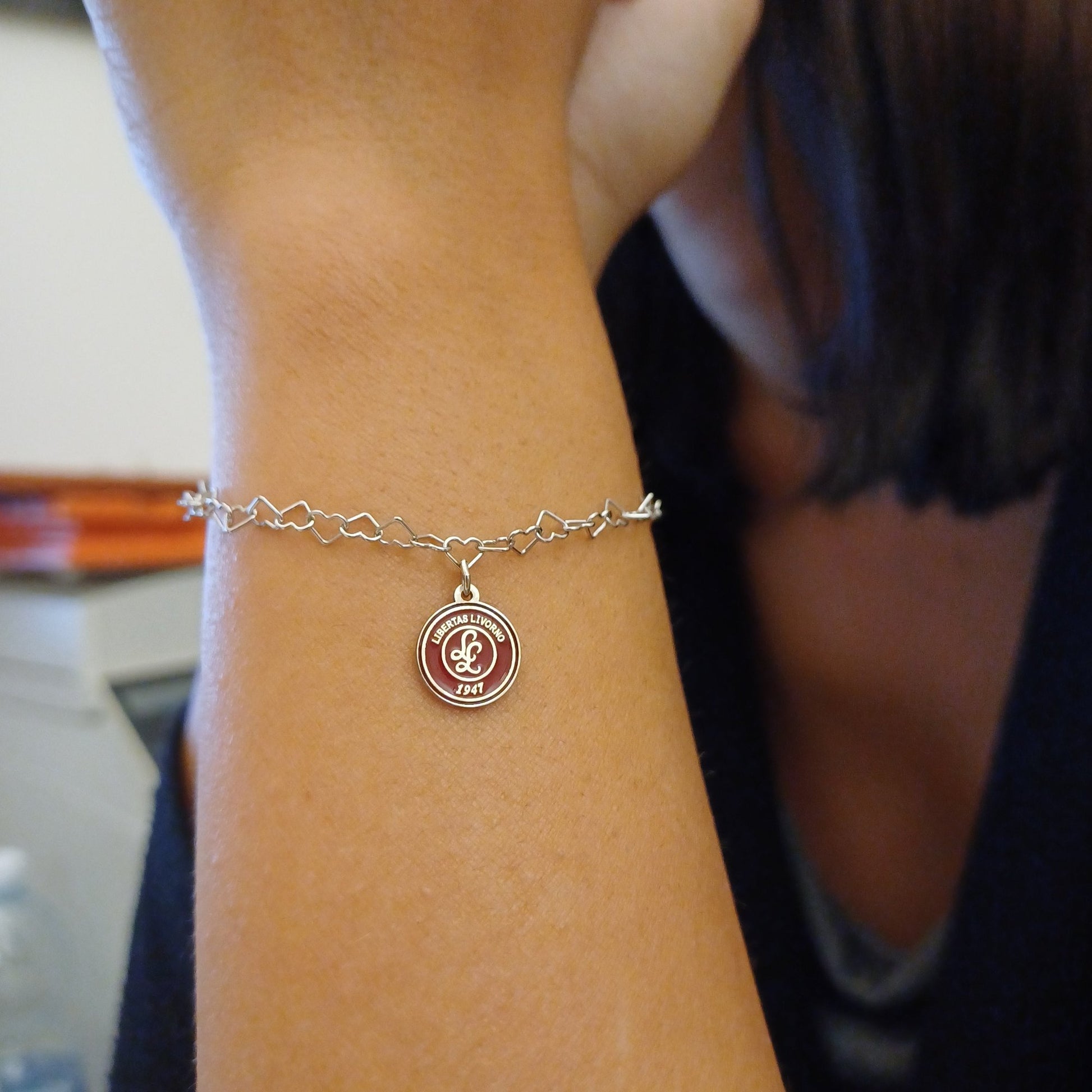 Questo braccialetto in argento 925 ha una catena di maglie a forma di cuore e una medaglietta rotonda da 10 mm. La superficie della medaglietta è lavorata artigianalmente nel nostro laboratorio orafo a Livorno e presenta il logo della Libertas Livorno 1947, inciso a laser e smaltato a caldo con un ricco colore amaranto. È un pezzo elegante e sportivo, pensato per celebrare la passione e l'appartenenza. Ideale per chi cerca gioielli personalizzati e distintivi.