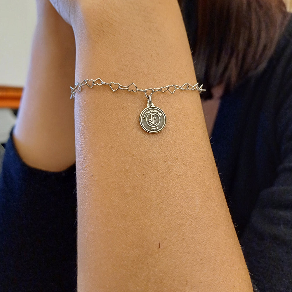 Questo braccialetto in argento 925 ha una catena di maglie a forma di cuore e una medaglietta rotonda da 10 mm. La superficie della medaglietta è lavorata artigianalmente nel nostro laboratorio orafo a Livorno e presenta il logo della Libertas Livorno 1947, inciso a laser e smaltato a caldo con un ricco colore amaranto. È un pezzo elegante e sportivo, pensato per celebrare la passione e l'appartenenza. Ideale per chi cerca gioielli personalizzati e distintivi.