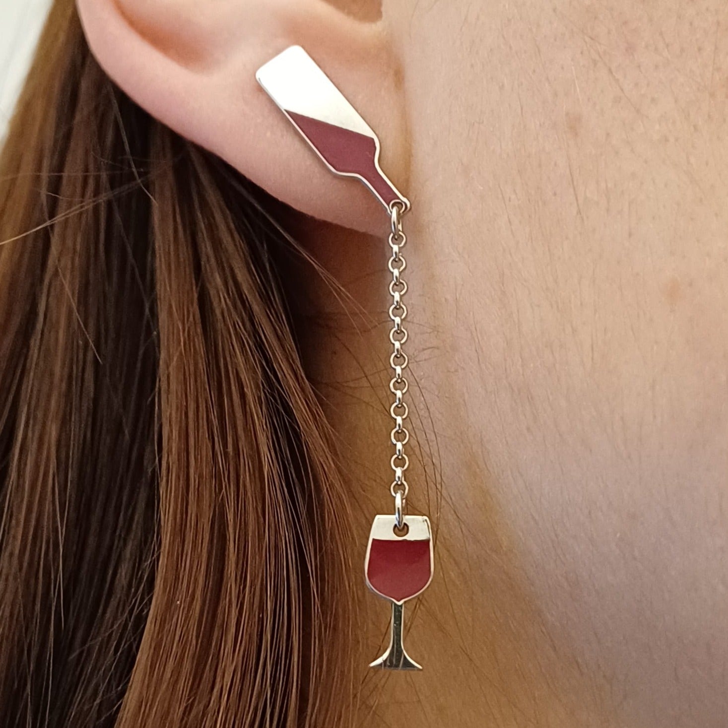 Questi orecchini pendenti in argento 925 sono composti da due elementi collegati da una sottile catenella. La parte superiore rappresenta il collo di una bottiglia, mentre il ciondolo inferiore raffigura un calice da vino inclinato, come se stesse versando il suo contenuto. Alcune versioni presentano una smaltatura a caldo in un intenso colore rosso vino. La superficie è incisa al laser, con dettagli ben definiti. Leggeri e lunghi 5 cm, si muovono delicatamente seguendo i movimenti del viso.