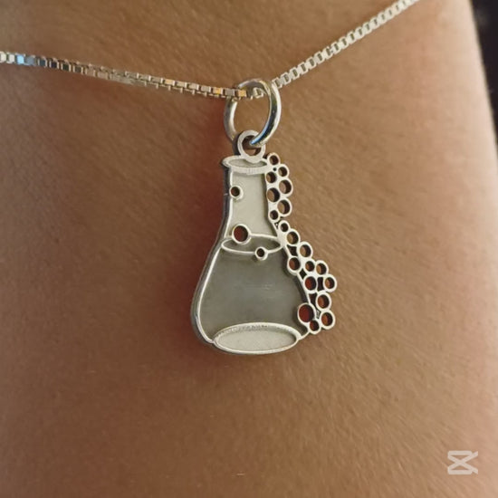 Il ciondolo rappresenta una piccola ampolla da laboratorio, alta circa 1,3 cm, realizzata in argento 925. La superficie è liscia e satinata, con rilievi che rappresentano delle bollicine che sembrano salire lungo il bordo. L’oggetto è leggero, con forme morbide e linee dolci. Al tatto, le bolle e il bordo dell’ampolla offrono una piacevole variazione tra pieno e vuoto, rendendolo facile da riconoscere e molto evocativo.