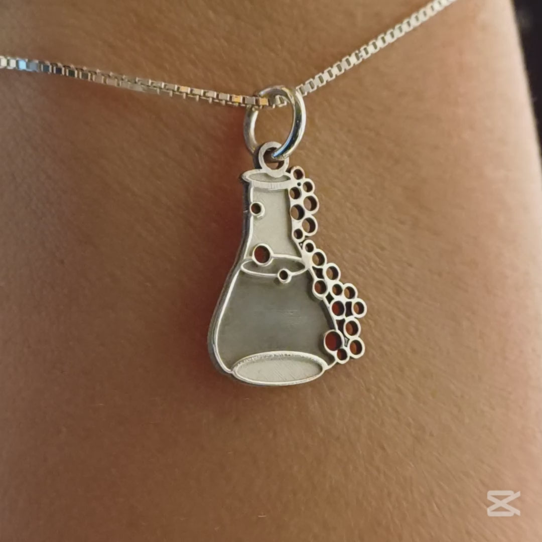 Il ciondolo rappresenta una piccola ampolla da laboratorio, alta circa 1,3 cm, realizzata in argento 925. La superficie è liscia e satinata, con rilievi che rappresentano delle bollicine che sembrano salire lungo il bordo. L’oggetto è leggero, con forme morbide e linee dolci. Al tatto, le bolle e il bordo dell’ampolla offrono una piacevole variazione tra pieno e vuoto, rendendolo facile da riconoscere e molto evocativo.