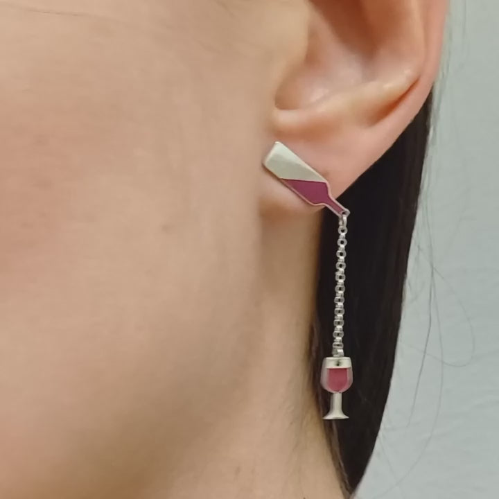 Questi orecchini pendenti in argento 925 sono composti da due elementi collegati da una sottile catenella. La parte superiore rappresenta il collo di una bottiglia, mentre il ciondolo inferiore raffigura un calice da vino inclinato, come se stesse versando il suo contenuto. Alcune versioni presentano una smaltatura a caldo in un intenso colore rosso vino. La superficie è incisa al laser, con dettagli ben definiti. Leggeri e lunghi 5 cm, si muovono delicatamente seguendo i movimenti del viso.