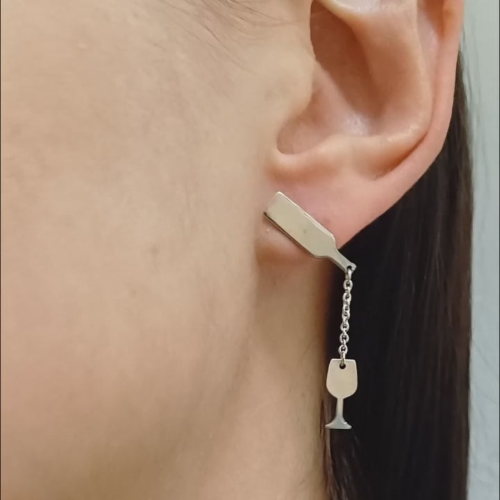 Questi orecchini pendenti in argento 925 sono composti da due elementi collegati da una sottile catenella. La parte superiore rappresenta il collo di una bottiglia, mentre il ciondolo inferiore raffigura un calice da vino inclinato, come se stesse versando il suo contenuto. Alcune versioni presentano una smaltatura a caldo in un intenso colore rosso vino. La superficie è incisa al laser, con dettagli ben definiti. Leggeri e lunghi 5 cm, si muovono delicatamente seguendo i movimenti del viso.